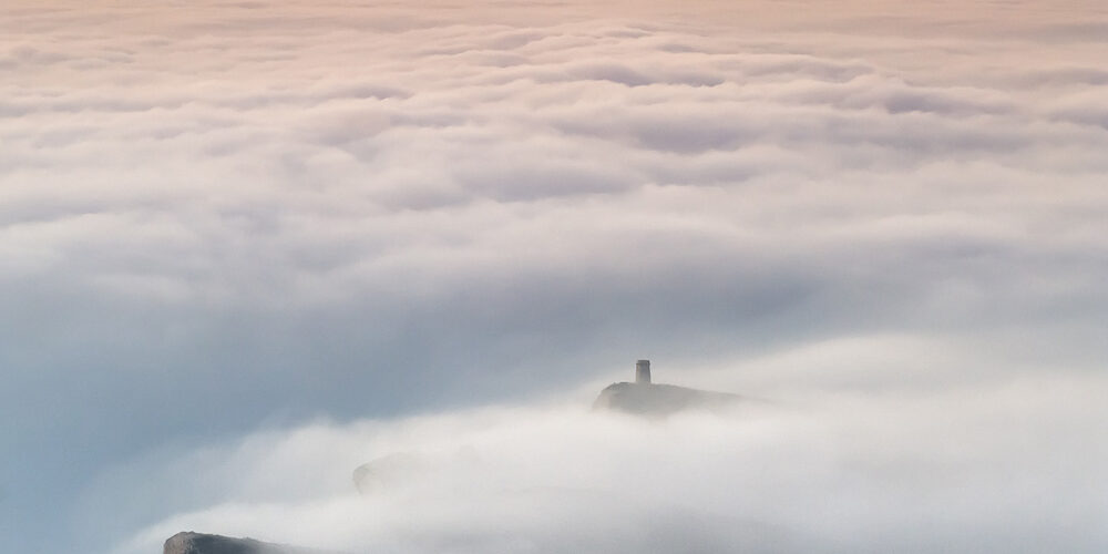 Fotografía de un amanecer sobre un mar de nubes, con acantilados y una torre destacándose entre la niebla. Los tonos cálidos en el cielo añaden un toque de serenidad y majestuosidad a la escena.