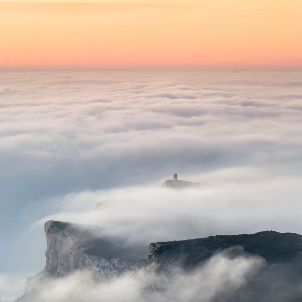 Fotografía de un amanecer sobre un mar de nubes, con acantilados y una torre destacándose entre la niebla. Los tonos cálidos en el cielo añaden un toque de serenidad y majestuosidad a la escena.