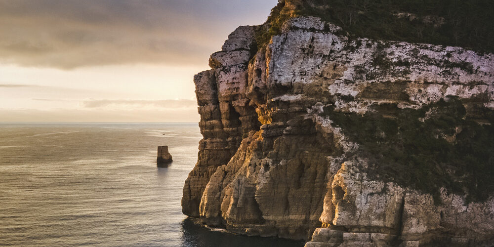 Fotografía de un acantilado iluminado por la luz dorada del amanecer, destacando la textura de las rocas y el azul profundo del mar Mediterráneo.