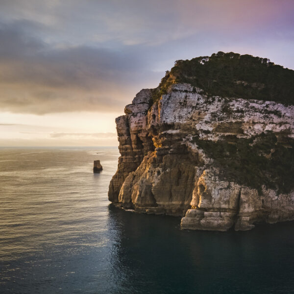 Fotografía de un acantilado iluminado por la luz dorada del amanecer, destacando la textura de las rocas y el azul profundo del mar Mediterráneo.