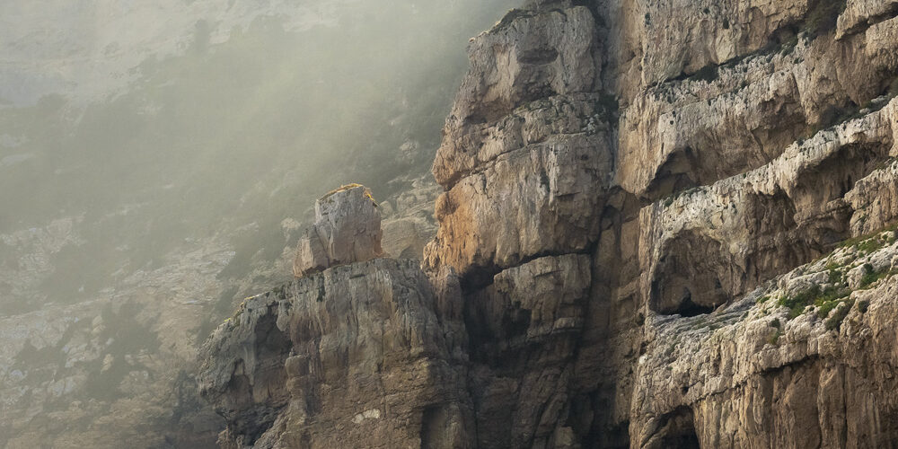 Detalle de un acantilado con rayos de luz filtrándose entre las rocas, creando un contraste dramático en el paisaje costero.