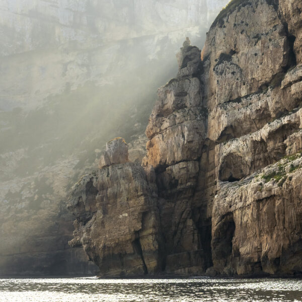 Detalle de un acantilado con rayos de luz filtrándose entre las rocas, creando un contraste dramático en el paisaje costero.