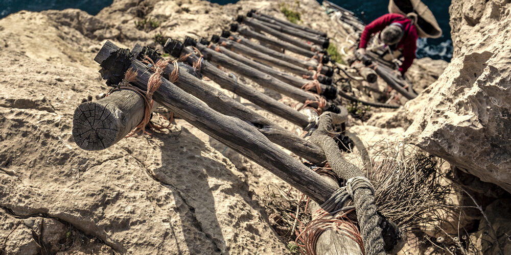 Vista de una escalera de madera anclada en un acantilado, descendiendo hacia el mar, enmarcando la dureza y belleza del entorno.