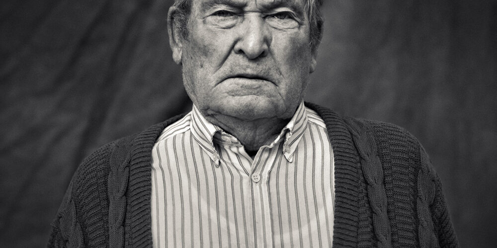Retrato en blanco y negro de un hombre mayor con jersey tejido y camisa a rayas, expresando seriedad. Fotografía artística para proyectos documentales.