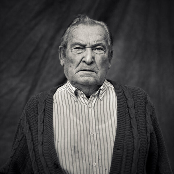 Retrato en blanco y negro de un hombre mayor con jersey tejido y camisa a rayas, expresando seriedad. Fotografía artística para proyectos documentales.