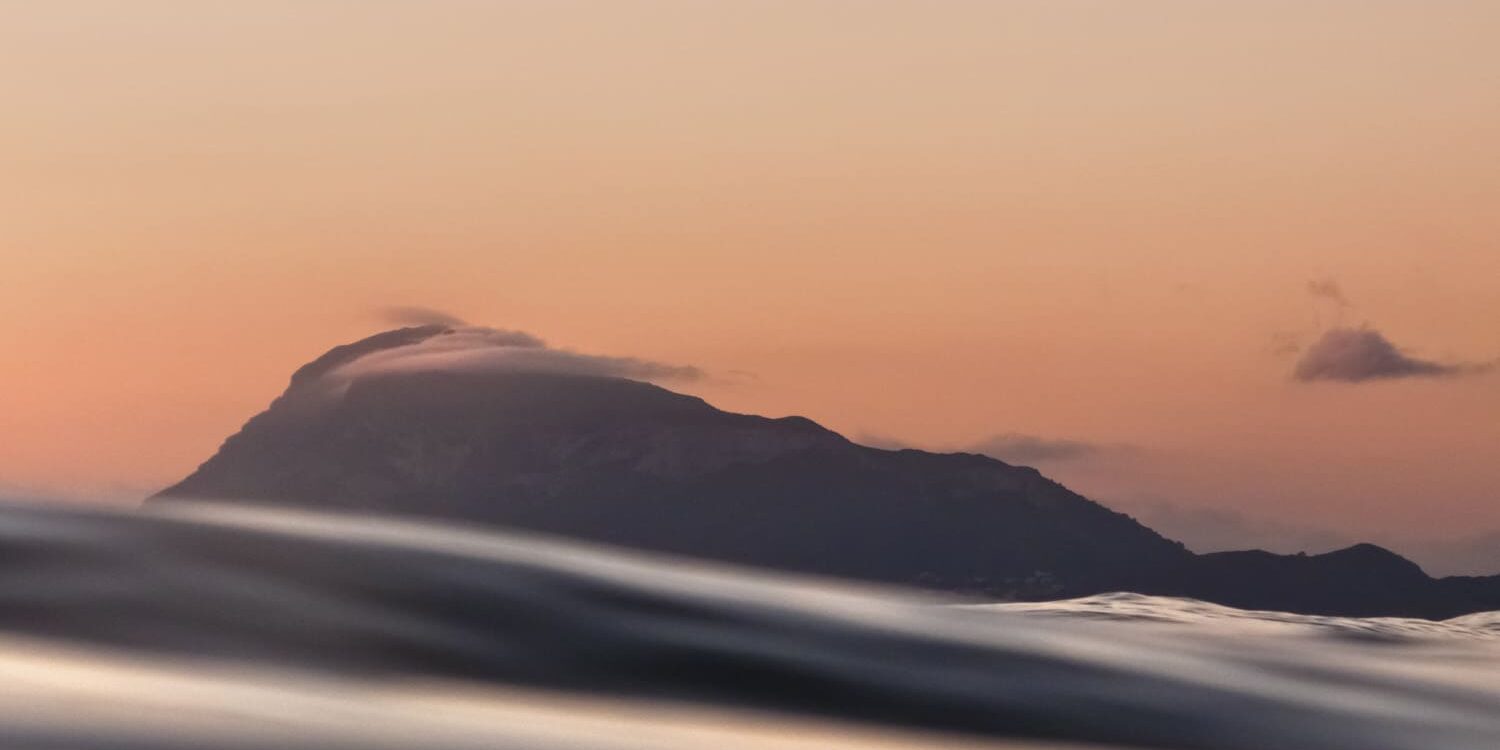 Fotografía del Montgó visto desde el nivel del mar al atardecer, con suaves tonos cálidos en el cielo y ondas en primer plano que crean una composición tranquila y elegante.