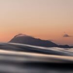 Fotografía del Montgó visto desde el nivel del mar al atardecer, con suaves tonos cálidos en el cielo y ondas en primer plano que crean una composición tranquila y elegante.