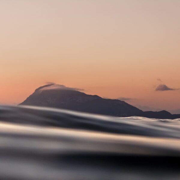 Fotografía del Montgó visto desde el nivel del mar al atardecer, con suaves tonos cálidos en el cielo y ondas en primer plano que crean una composición tranquila y elegante.