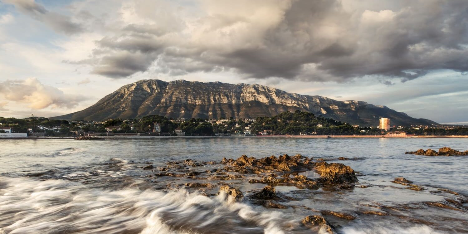 Fotografía del Montgó desde la costa, con rocas en primer plano y el agua del mar en movimiento, bajo un cielo dramático lleno de nubes. Una escena que combina la fuerza del paisaje marino y la majestuosidad de la montaña.