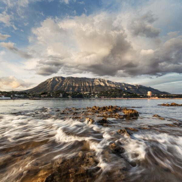 Fotografía del Montgó desde la costa, con rocas en primer plano y el agua del mar en movimiento, bajo un cielo dramático lleno de nubes. Una escena que combina la fuerza del paisaje marino y la majestuosidad de la montaña.