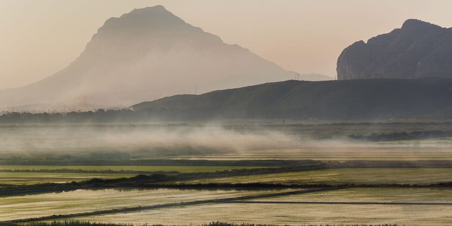 Fotografía de un paisaje rural con campos inundados al amanecer, creando reflejos sutiles bajo una ligera niebla. Al fondo, la imponente silueta del Montgó enmarca la escena, mientras una figura solitaria camina en la distancia. Una composición que transmite calma y conexión con la naturaleza.