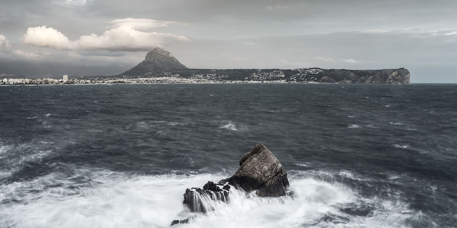 Fotografía del Montgó y la costa de Jávea en un día nublado, con el mar agitado y olas rompiendo contra las rocas en primer plano. Una escena que captura la fuerza y la belleza del paisaje marino.