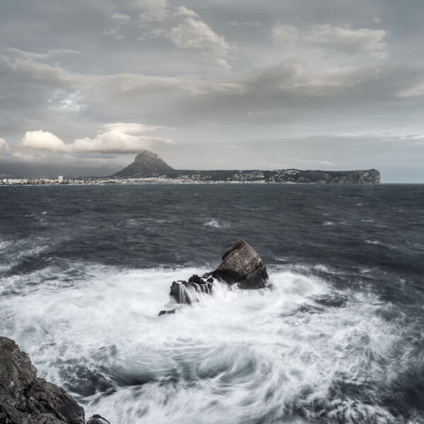 Fotografía del Montgó y la costa de Jávea en un día nublado, con el mar agitado y olas rompiendo contra las rocas en primer plano. Una escena que captura la fuerza y la belleza del paisaje marino.