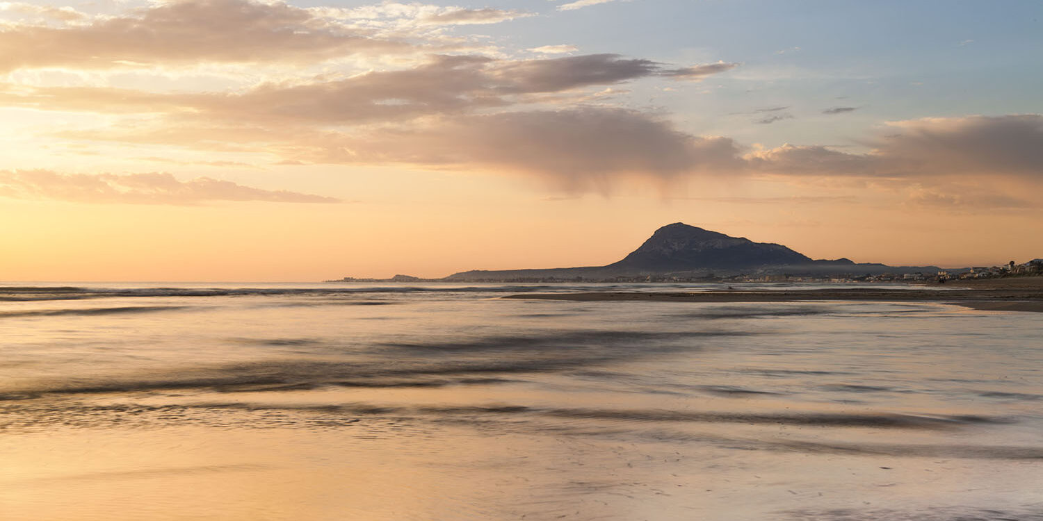 Fotografía de la playa al atardecer con el Montgó en el horizonte, reflejando tonos dorados y cálidos sobre el agua tranquila. Una escena serena que combina la belleza de la costa y la montaña.