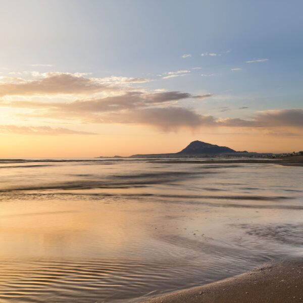 Fotografía de la playa al atardecer con el Montgó en el horizonte, reflejando tonos dorados y cálidos sobre el agua tranquila. Una escena serena que combina la belleza de la costa y la montaña.
