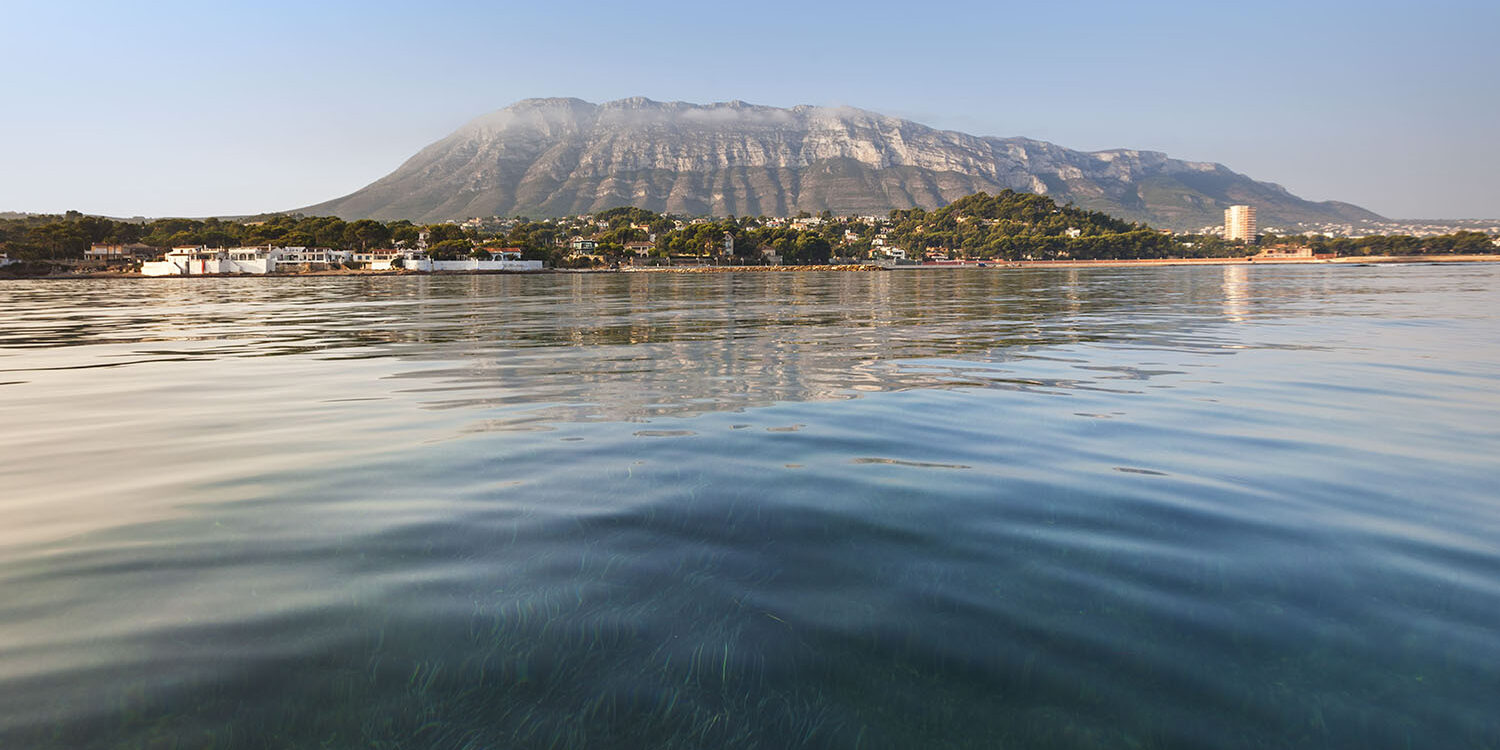 Fotografía del Montgó visto desde el agua, con la costa de Jávea en el horizonte y el fondo marino visible a través del agua cristalina. Una composición que combina la majestuosidad de la montaña y la claridad del entorno marino.