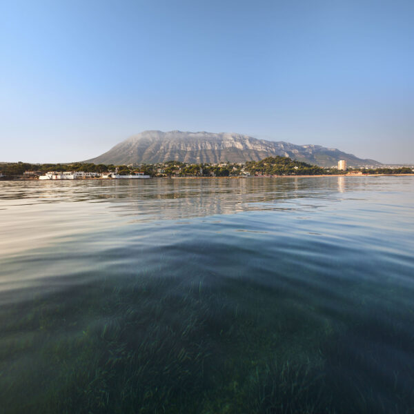 Fotografía del Montgó visto desde el agua, con la costa de Jávea en el horizonte y el fondo marino visible a través del agua cristalina. Una composición que combina la majestuosidad de la montaña y la claridad del entorno marino.