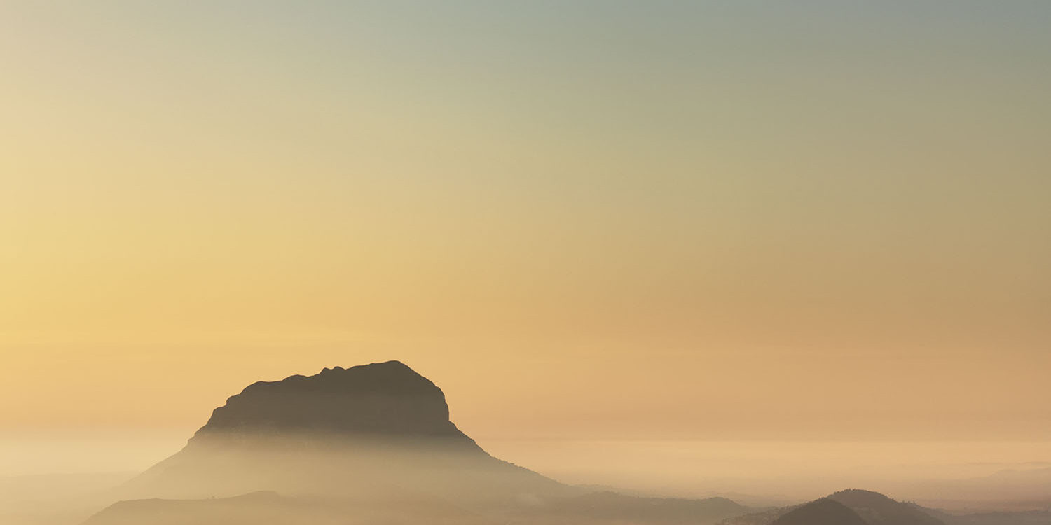 Fotografía del Montgó emergiendo entre una suave niebla al amanecer, con tonos dorados y cálidos que iluminan el horizonte. Una escena que transmite tranquilidad y la majestuosidad del paisaje natural.