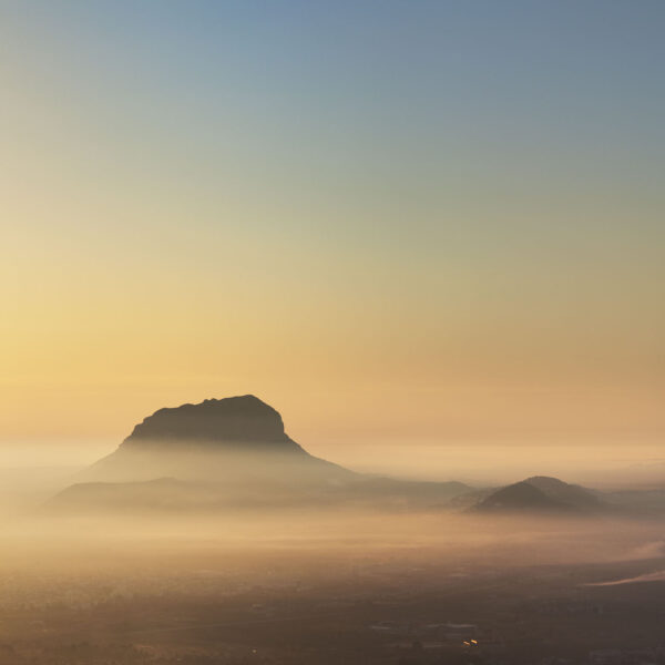 Fotografía del Montgó emergiendo entre una suave niebla al amanecer, con tonos dorados y cálidos que iluminan el horizonte. Una escena que transmite tranquilidad y la majestuosidad del paisaje natural.