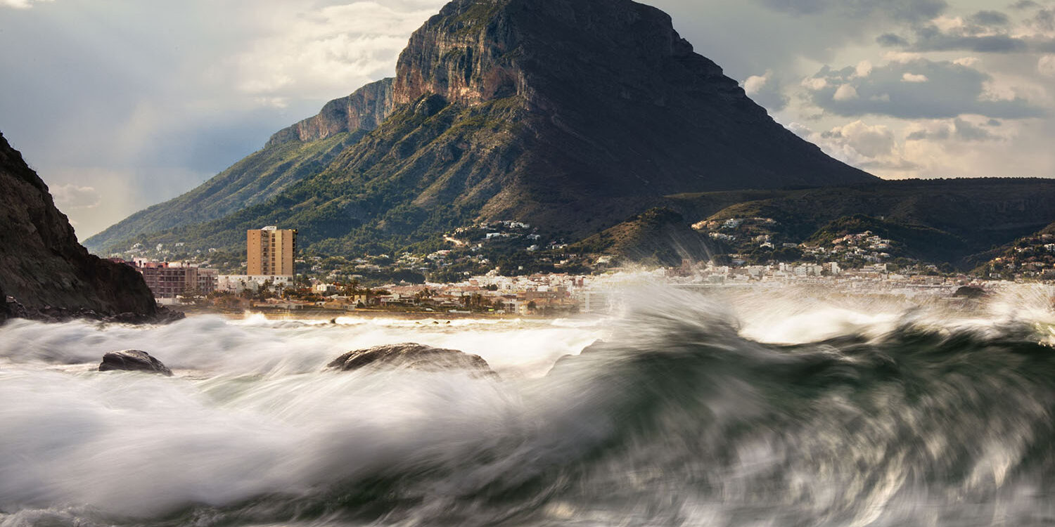 Fotografía del Montgó desde la costa, con olas enérgicas rompiendo en primer plano bajo un cielo parcialmente nublado. Una escena que captura la fuerza del mar y la majestuosidad de la montaña en un entorno dinámico y natural.