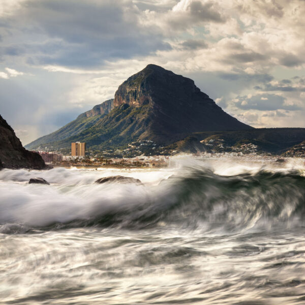 Fotografía del Montgó desde la costa, con olas enérgicas rompiendo en primer plano bajo un cielo parcialmente nublado. Una escena que captura la fuerza del mar y la majestuosidad de la montaña en un entorno dinámico y natural.