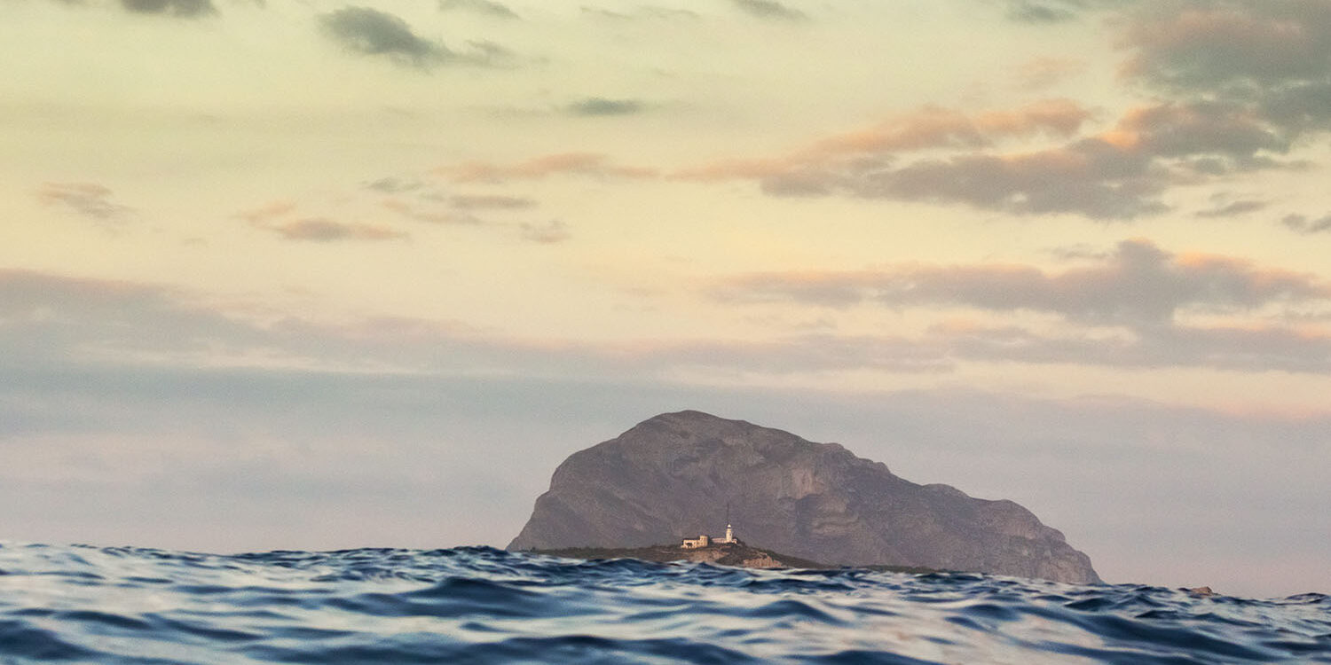 Fotografía del Montgó visto desde el nivel del mar al atardecer, con un cielo suavemente teñido de tonos pastel y el agua en movimiento en primer plano. Un faro se destaca en la distancia, añadiendo un toque de serenidad a la escena.