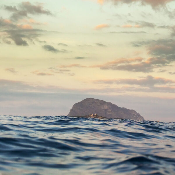 Fotografía del Montgó visto desde el nivel del mar al atardecer, con un cielo suavemente teñido de tonos pastel y el agua en movimiento en primer plano. Un faro se destaca en la distancia, añadiendo un toque de serenidad a la escena.